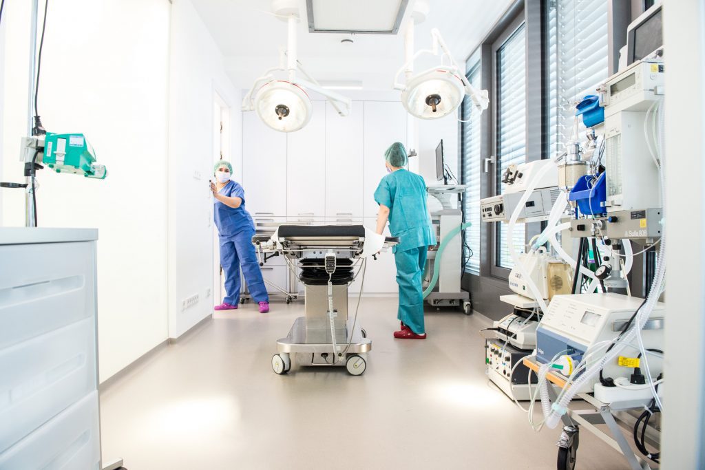 Zwei medizinische Fachkräfte in OP-Kitteln bereiten einen Operationssaal vor. Einer richtet die Geräte ein, der andere steht neben der Tür. Der Raum ist hell, mit verschiedenen medizinischen Geräten und Lampen ausgestattet und bereit für Eingriffe wie die Proktologie.