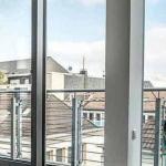 Große Glasfenster bieten einen Blick auf die Dächer der Stadt und den Turm einer Kathedrale in der Ferne. Draußen gibt es einen Balkon mit Geländer. Drinnen hängt ein kleines Poster mit dem Gesicht einer lächelnden Person und einer Werbung für Laser-Hautbehandlungen.