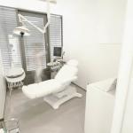 Ein modernes Zahnarztzimmer mit Zahnarztstuhl, Deckenleuchte, Monitor und verschiedenen modernen Zahnarztinstrumenten. Das Zimmer verfügt über große Fenster mit Jalousien und weiße Wände, die für eine helle und einladende Umgebung sorgen. Den Patienten stehen auch moderne Technologien wie Laserbehandlungen zur Verfügung.