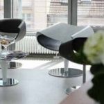 Ein moderner Wartebereich mit drei eleganten, schwarz-grauen Stühlen, einem durchsichtigen Glastisch mit Zeitschriften und großen Fenstern, die den Raum mit natürlichem Licht füllen. Im Vordergrund ist teilweise eine Pflanze zu sehen, die der ruhigen Umgebung, die sich ideal für eine Gesichtsberatung eignet, einen Hauch von Grün verleiht.